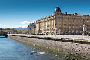 A large, historic building with a prominent dome and multiple balconies, situated beside a river with a stone embankment.