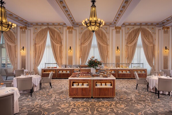 A luxurious hotel dining area with ornate chandeliers, elegant drapery, and a central buffet station.