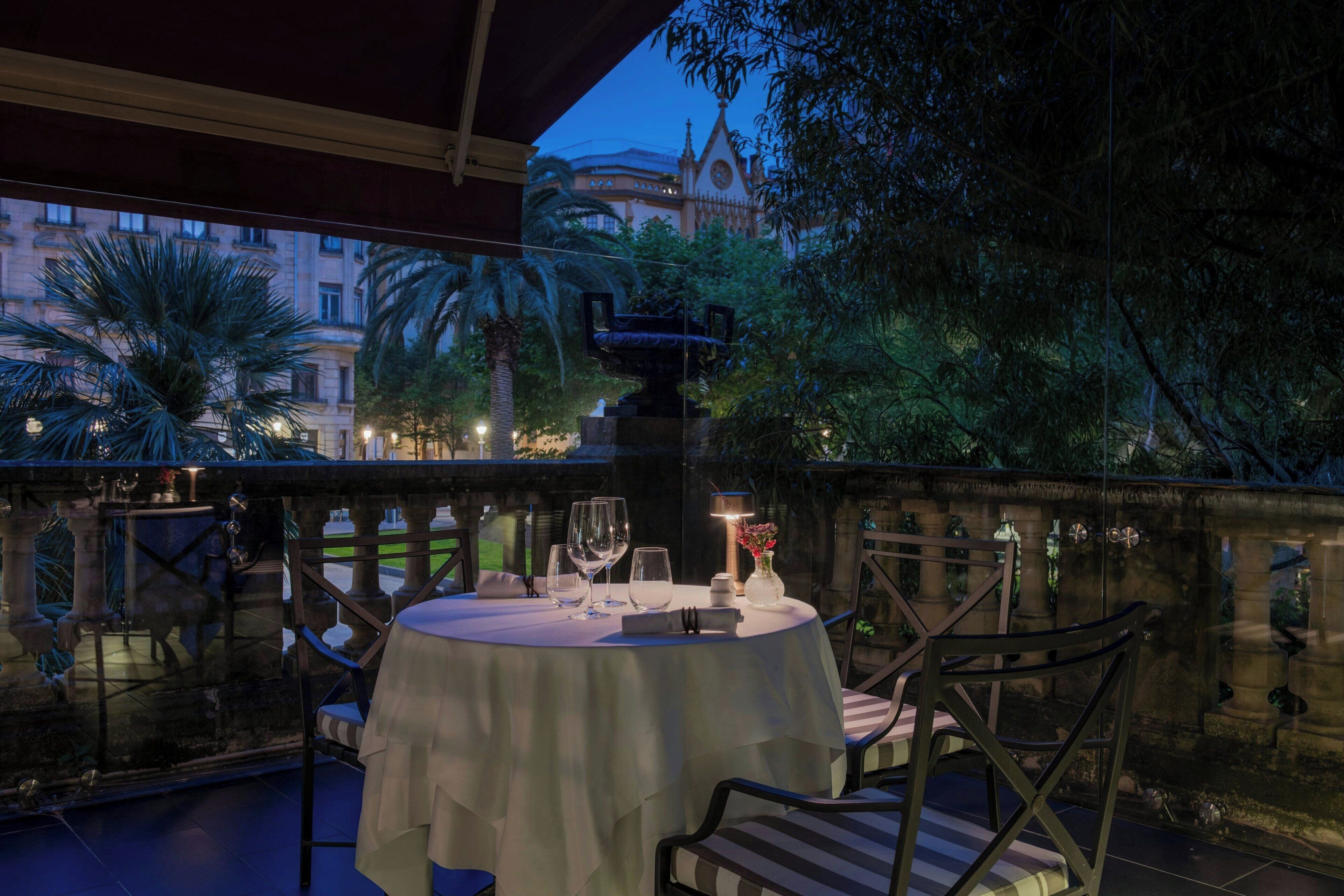 An outdoor dining area with a table set for two, surrounded by chairs, and a decorative fountain in the background.