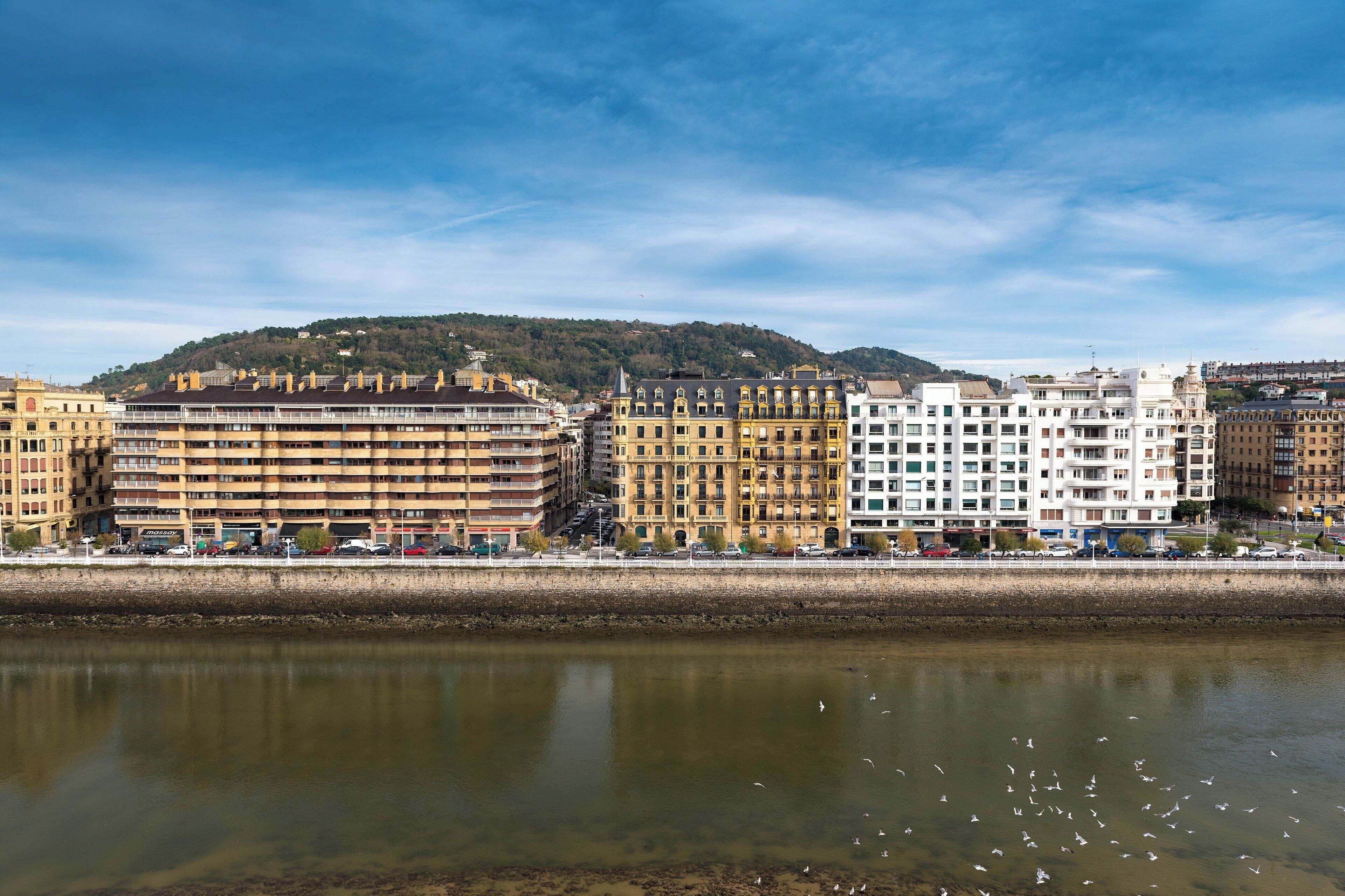 Un paisaje urbano frente al río, con edificios a lo largo de la ribera, una colina al fondo y cielo despejado.