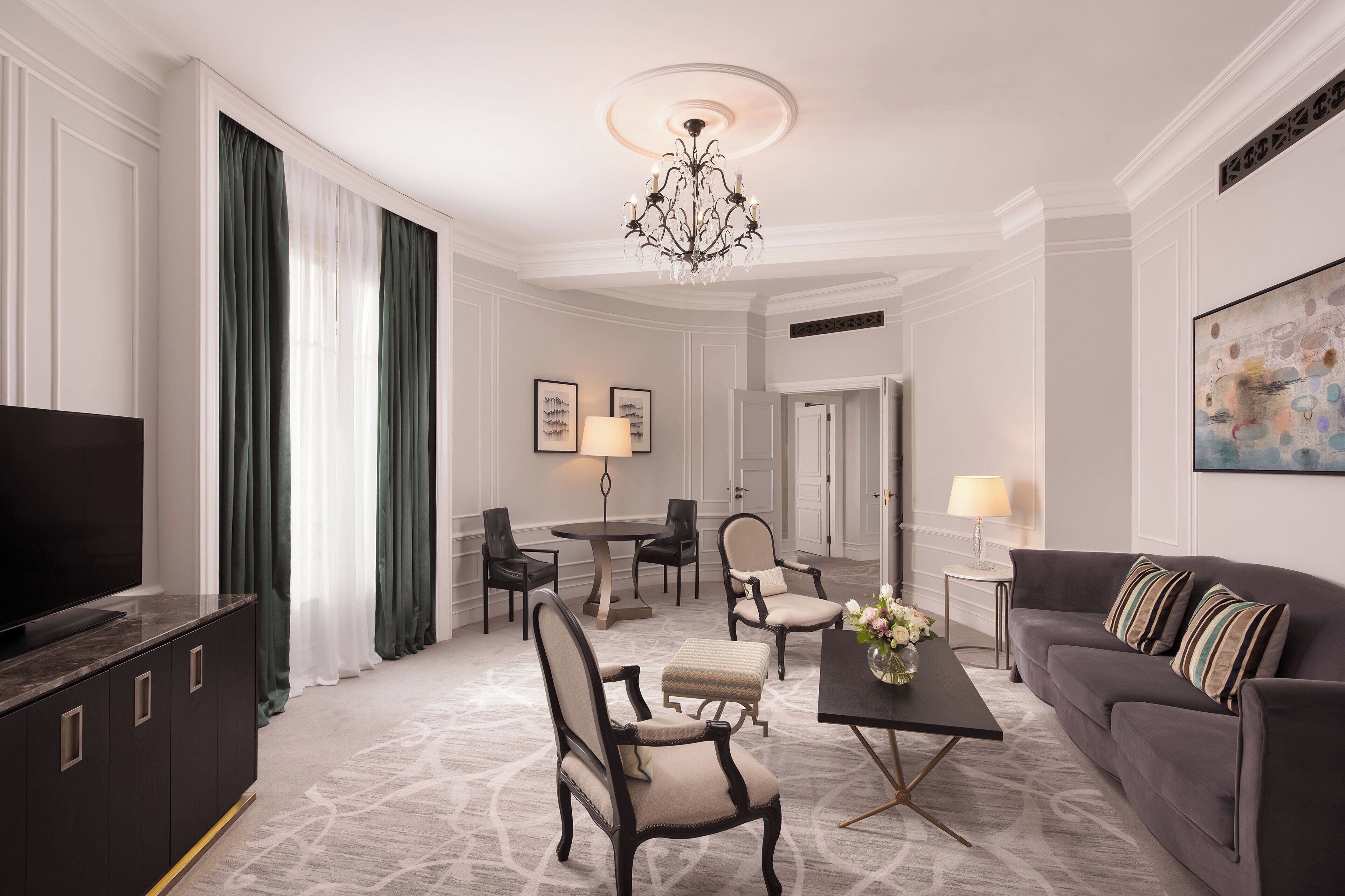 Una habitación de hotel moderna con un televisor de pantalla plana grande, un mueble de madera oscura, un sofá gris con cojines a rayas, una mesa de comedor redonda con sillas y una araña.