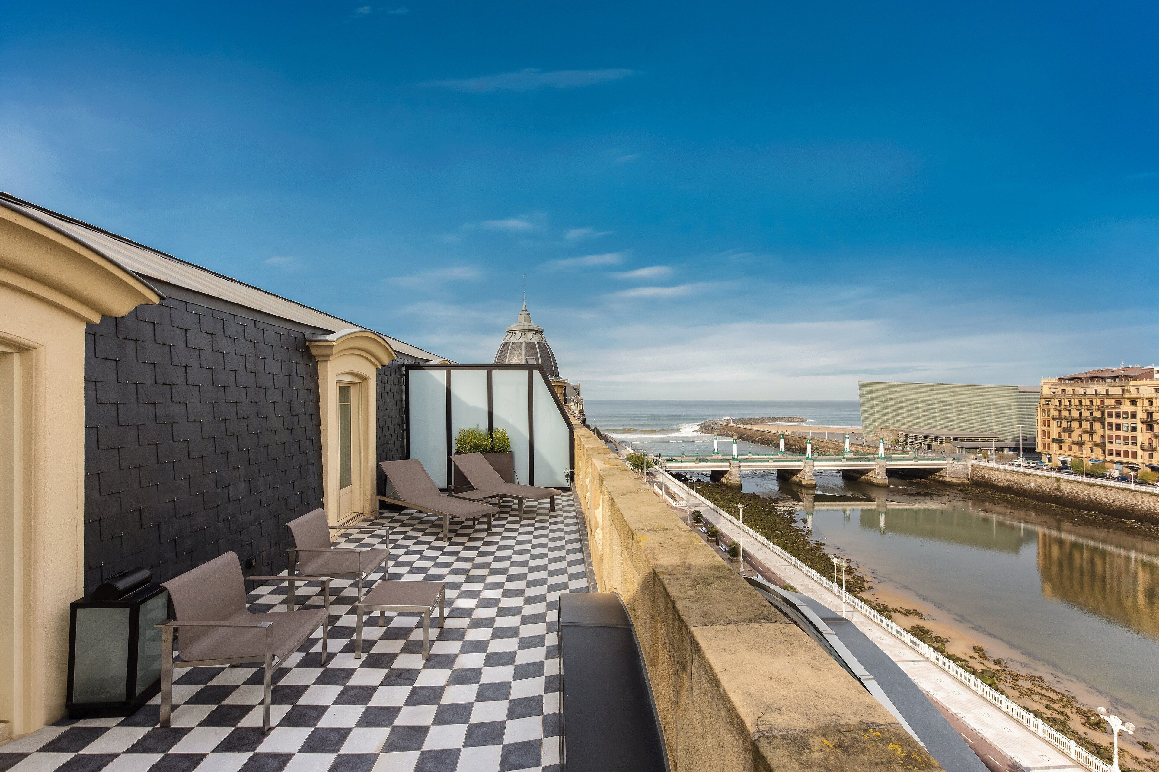A rooftop terrace with a checkered floor, lounge chairs, and a view of the sea and cityscape.