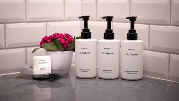 Cuatro productos capilares BYREDO Le Chemin sobre una superficie reflectante junto a una planta en maceta y una pastilla de jabón.