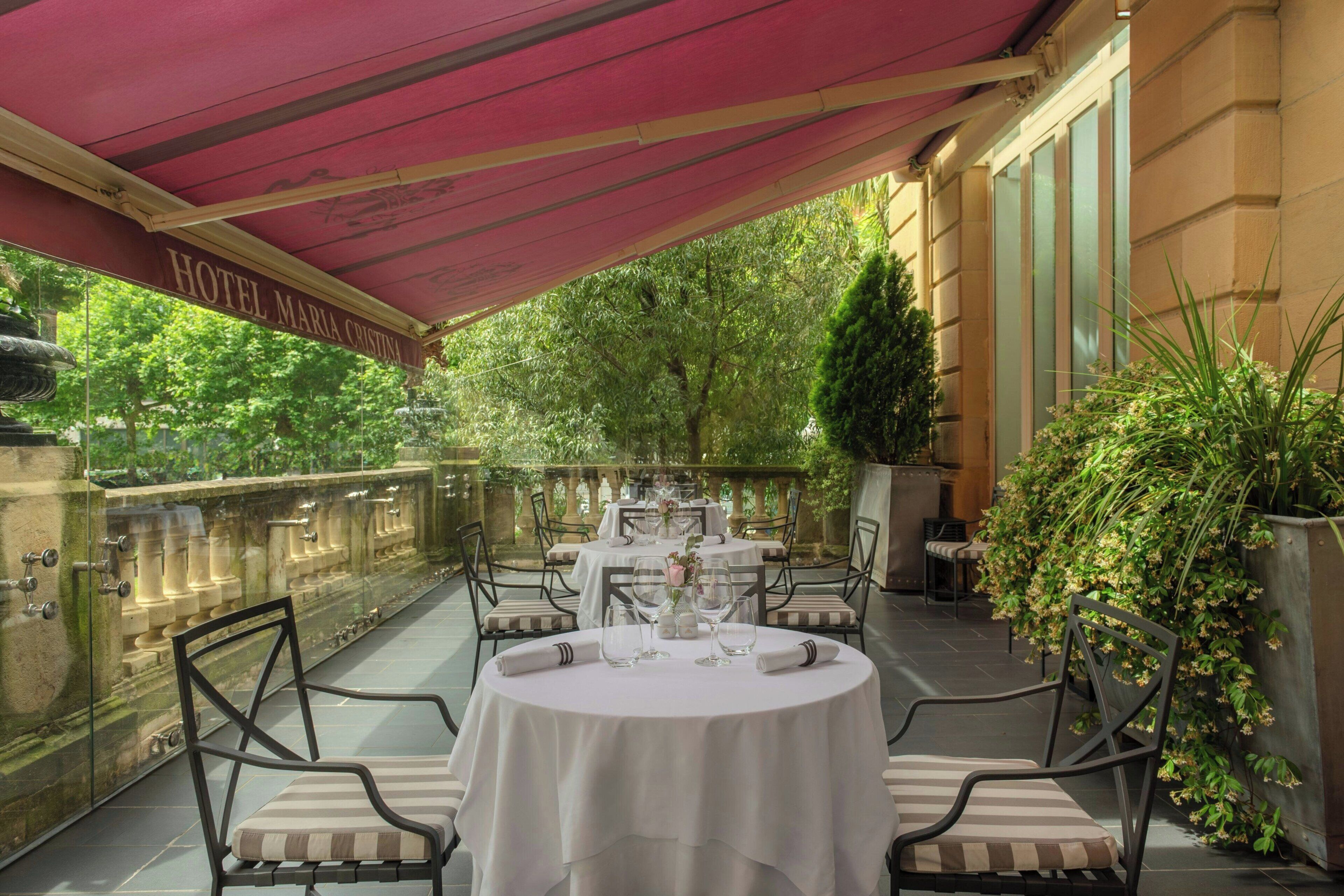 A covered outdoor dining area with round tables and chairs, a glass balustrade, and lush greenery in the background.