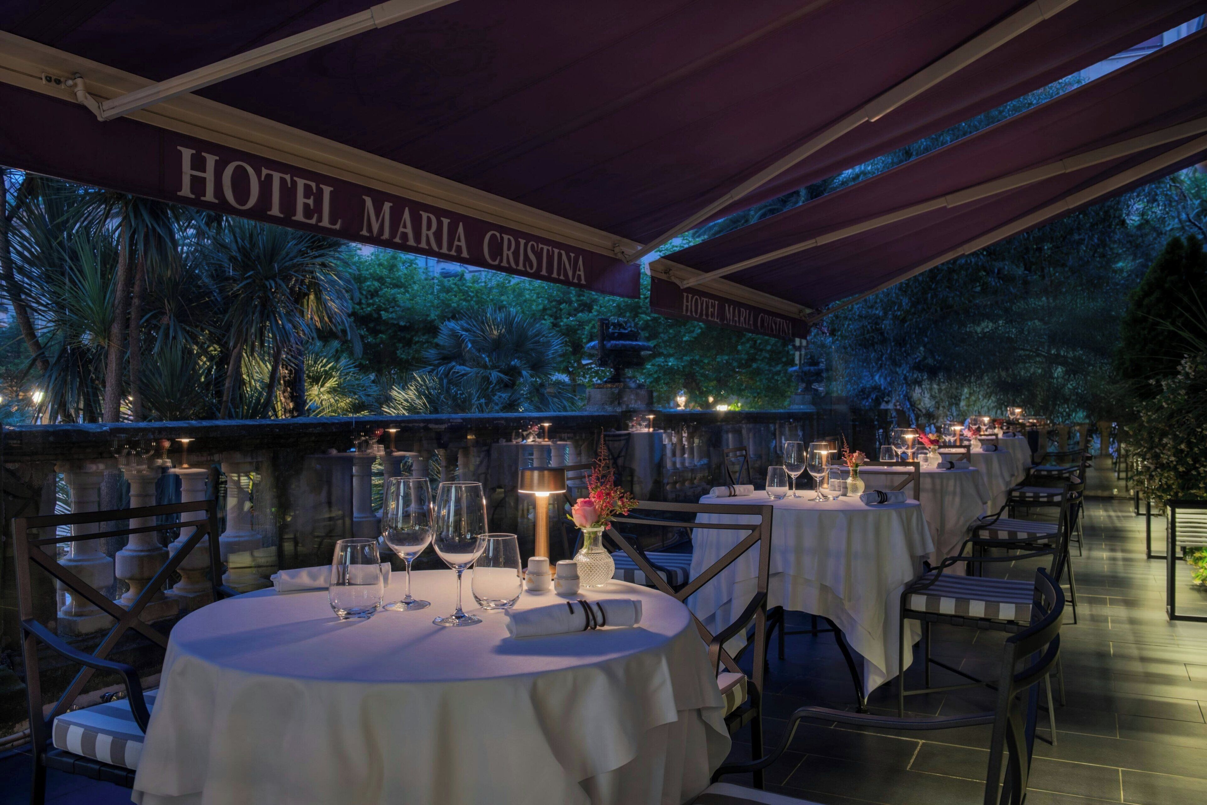 An outdoor dining area of Hotel Maria Cristina with round tables, chairs, and a stone balustrade.
