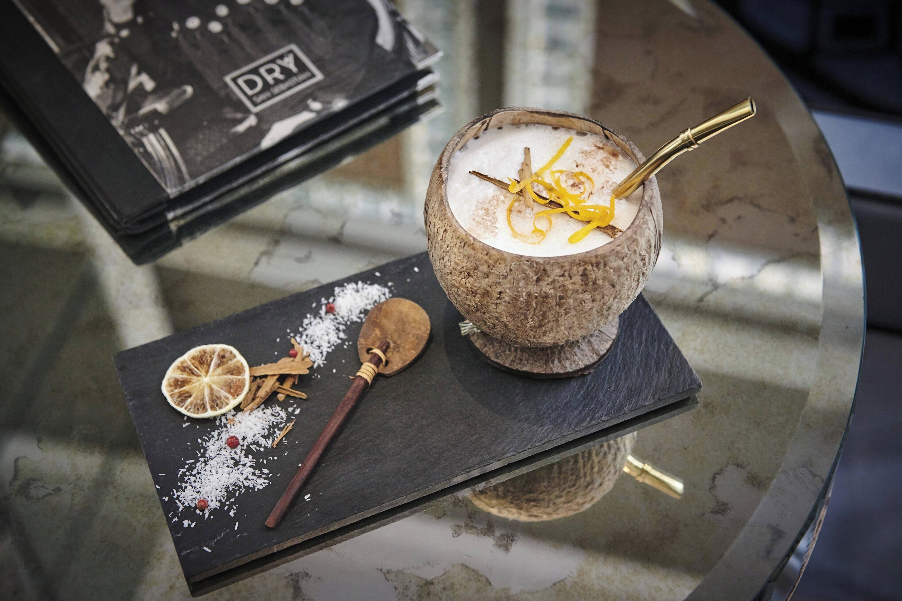 Bebida de coco sobre una mesa de vidrio, con pajita, adornada con ralladura de cítricos y una rama de canela.