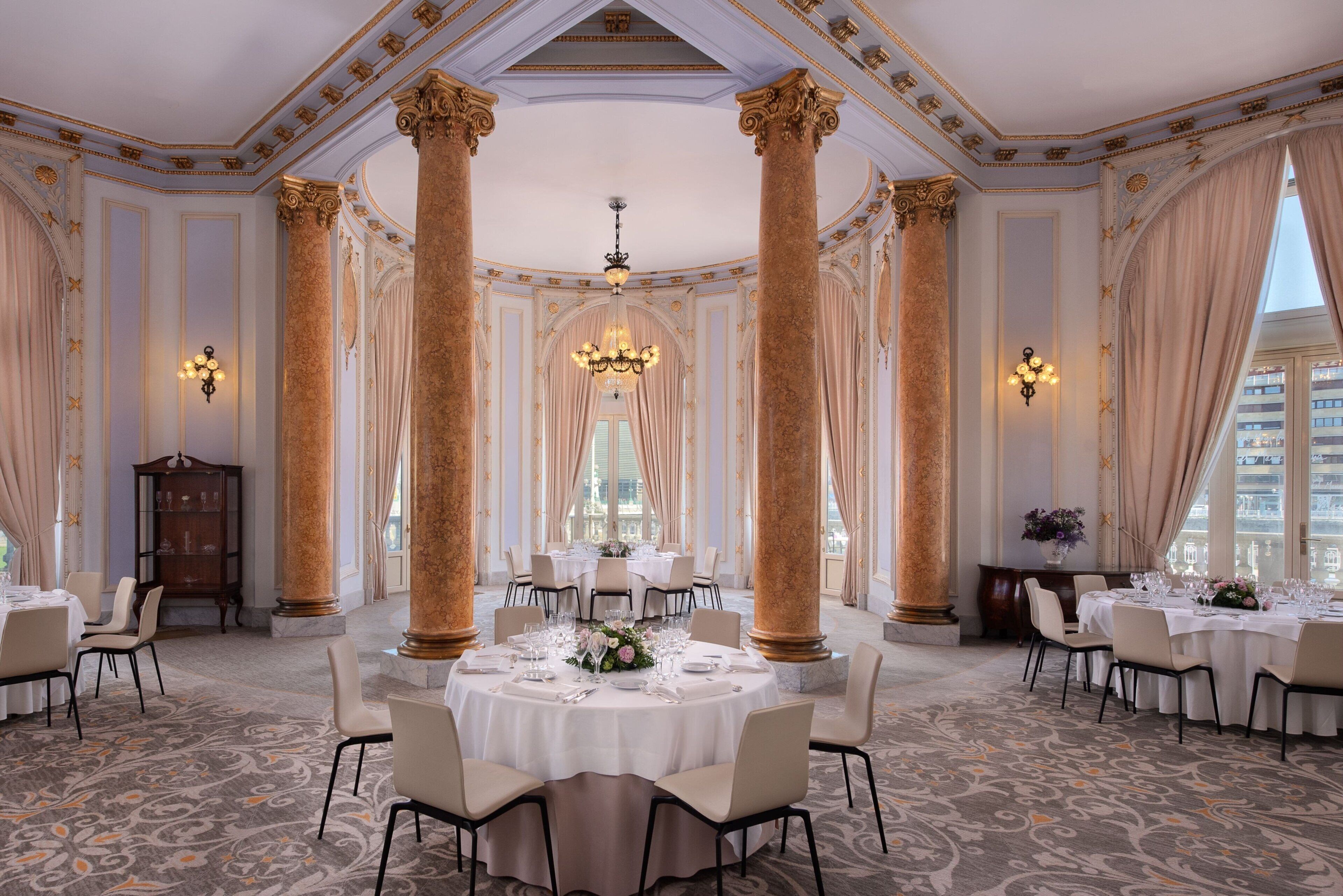 Un comedor amplio y elegante, con techos altos, columnas decorativas, candelabros y mesas redondas dispuestas para una comida.