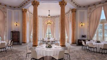 Un comedor amplio y elegante, con techos altos, columnas decorativas, candelabros y mesas redondas dispuestas para una comida.