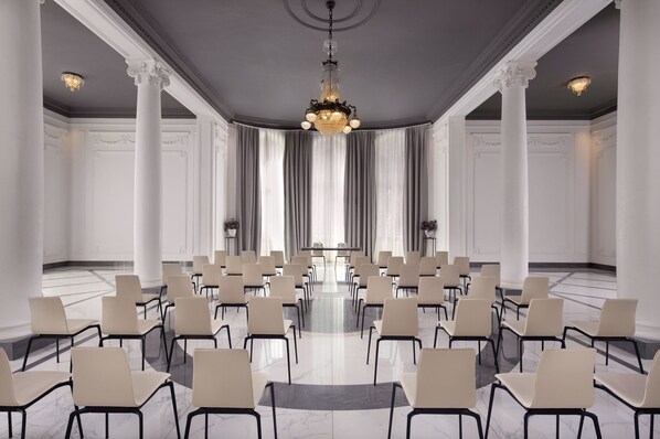 A conference room with rows of chairs, a chandelier, and large windows with curtains.