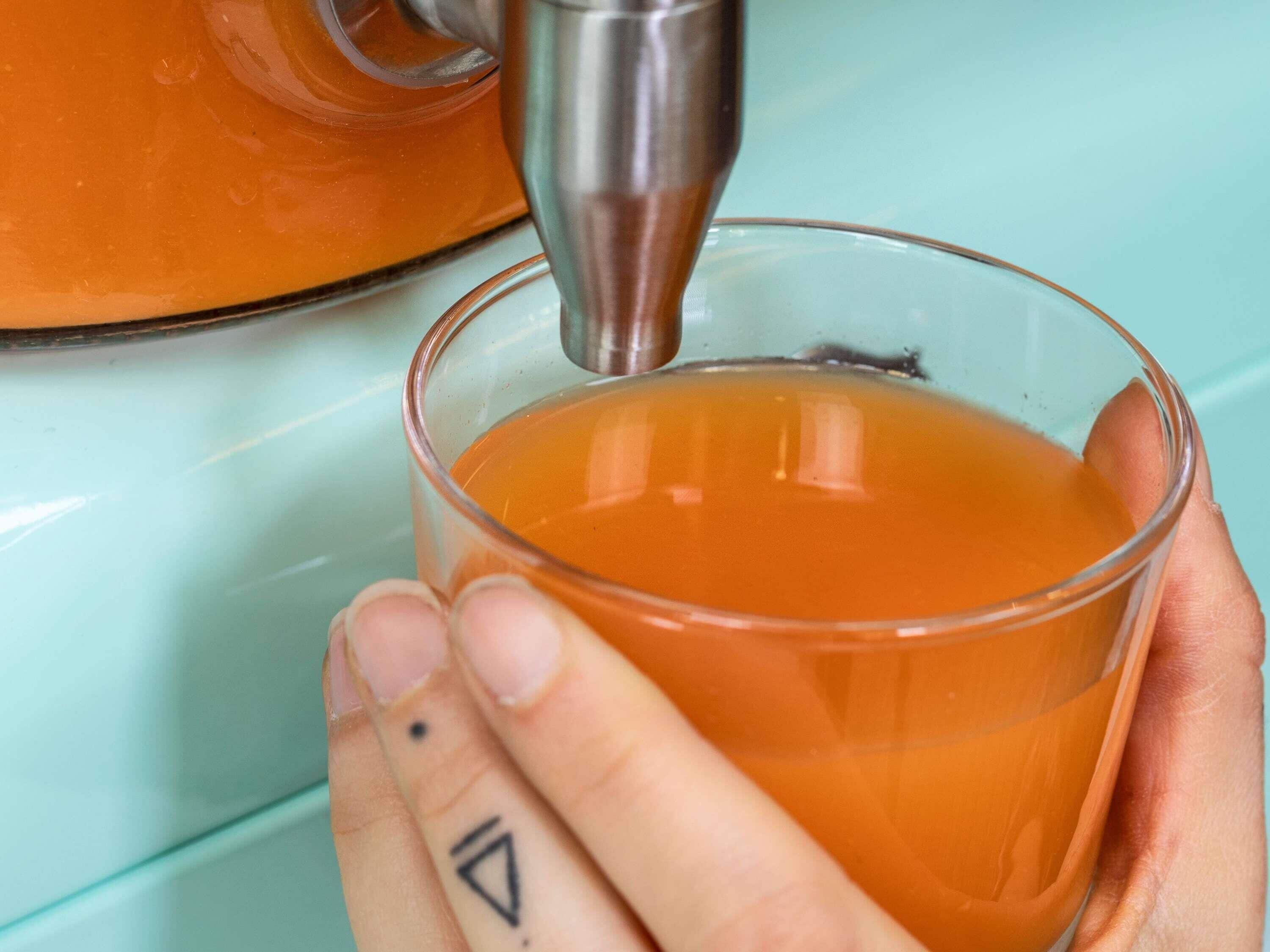 A hand holding a glass with a liquid, possibly tea, being poured from a metallic container.