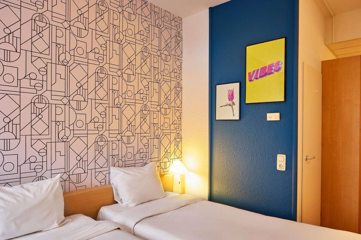 A hotel room with a geometric patterned wallpaper, a bed with white linens, a blue accent wall, and a framed picture with the word 'VIBES'.