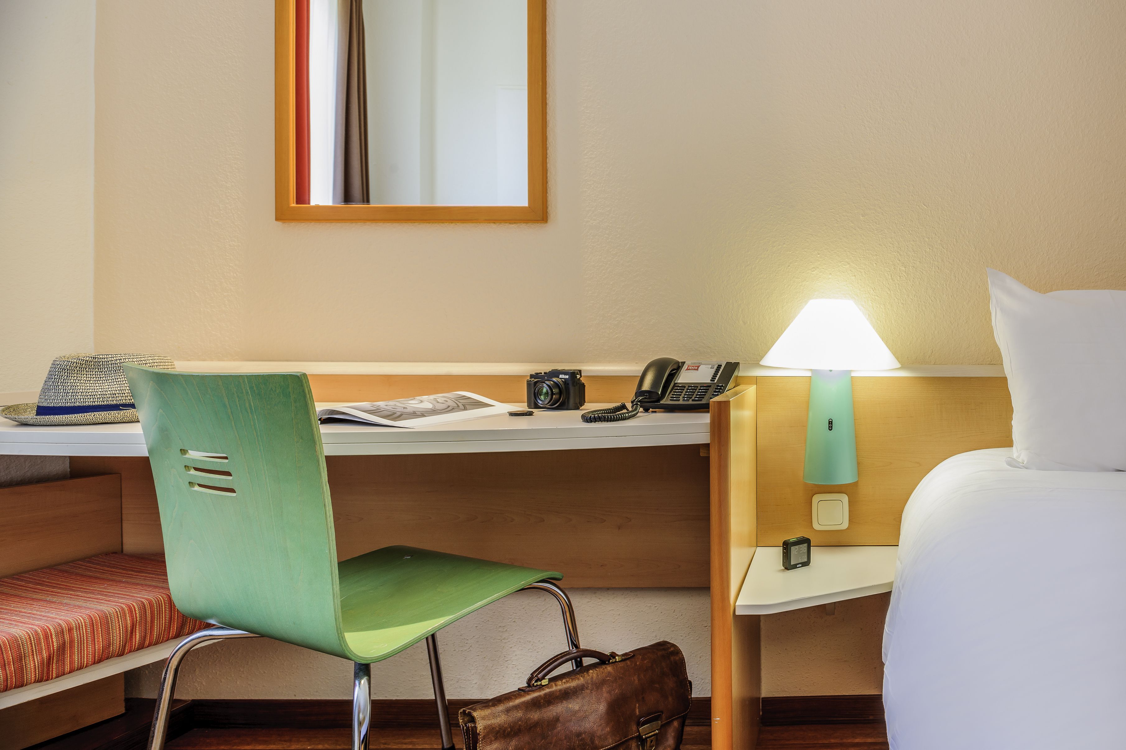 A hotel room with a bed, a desk with a lamp, a chair, a handbag, and a small table with a phone and a camera.