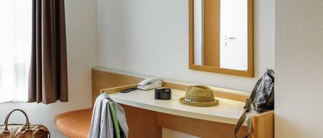 A hotel room with a wooden desk, a green chair, a bed, a window with curtains, a mirror, a telephone, a camera, a hat, and a handbag.