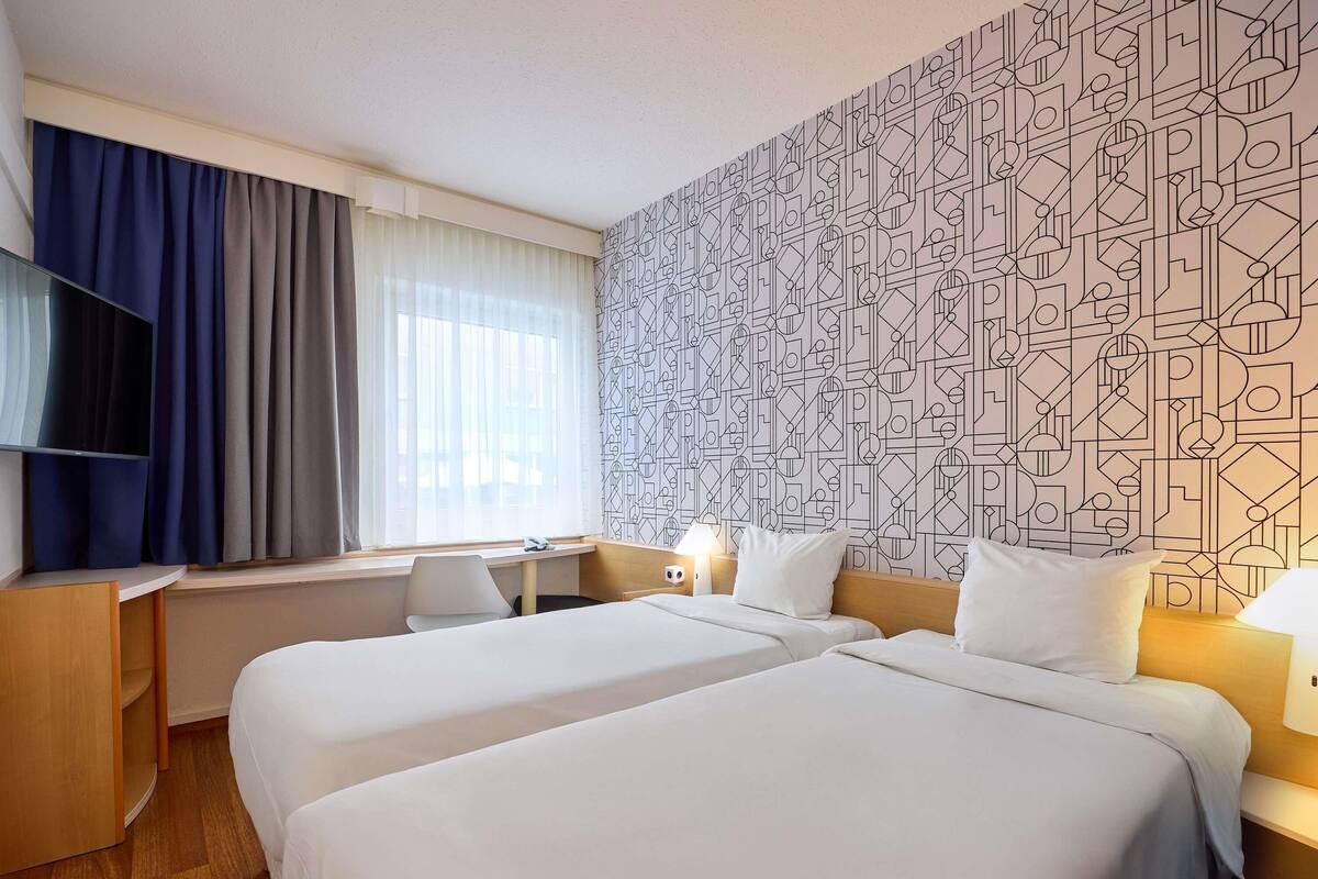 Habitación de hotel con dos camas, un escritorio, un televisor y un papel tapiz estampado.