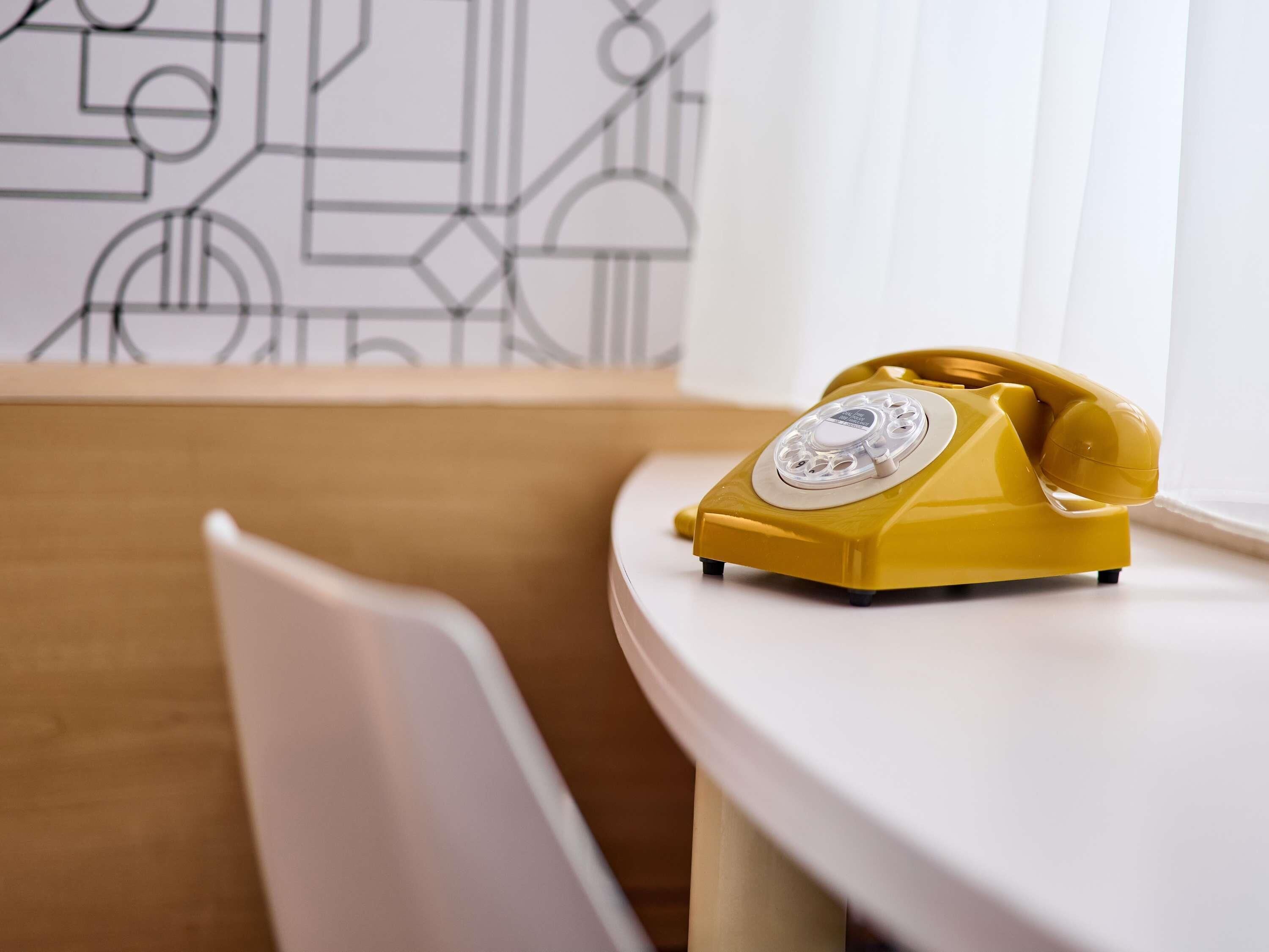 Ein gelbes Wählscheibentelefon steht auf einem weißen Tisch in einem Raum mit einer Wand mit geometrischem Muster.