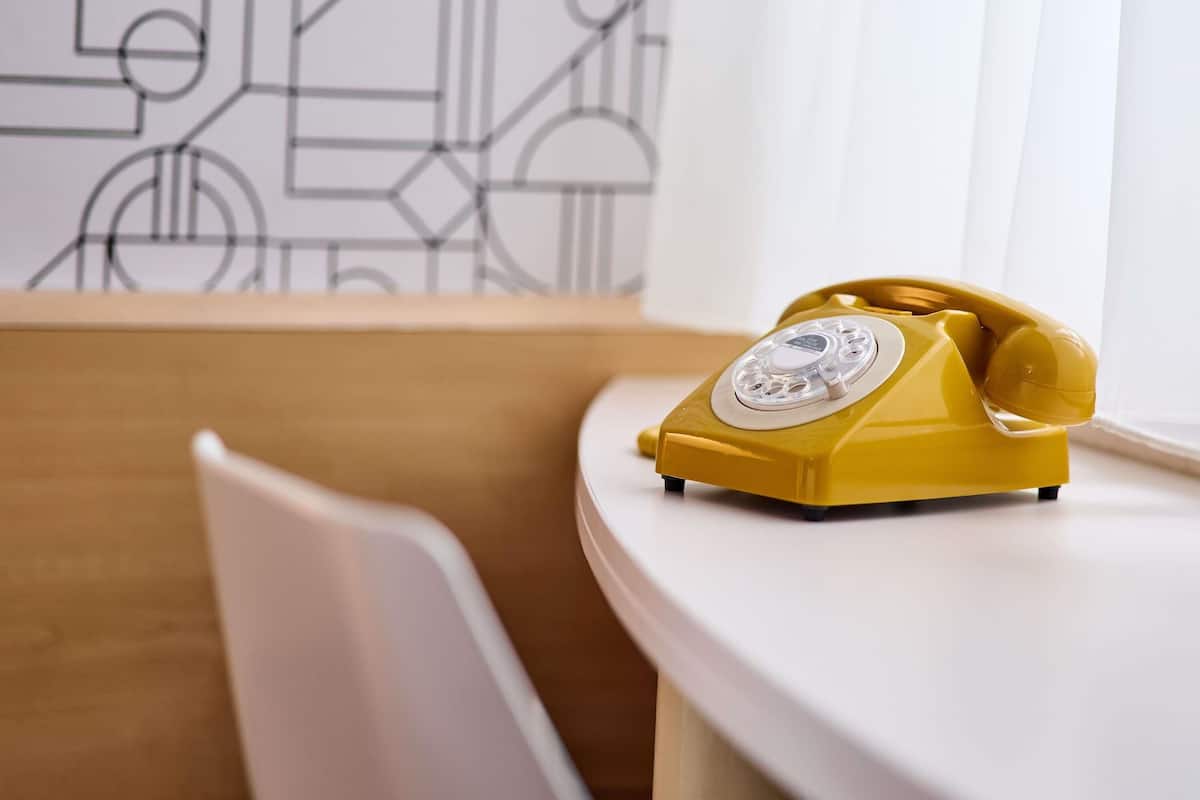 Un téléphone à cadran jaune est posé sur une table blanche dans une pièce dont le mur est décoré d’un motif géométrique.
