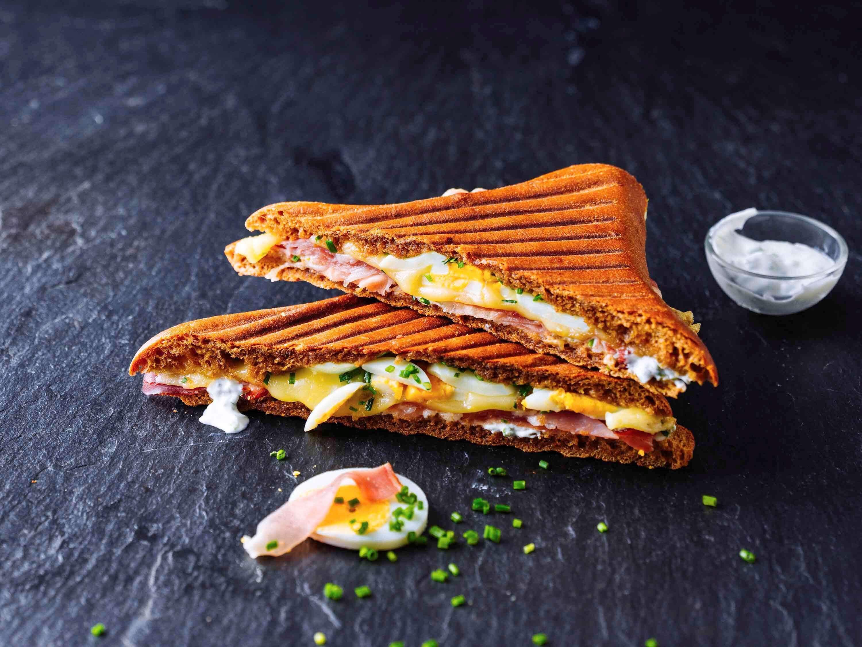 A sandwich cut in half, with visible layers of ham, cheese, and egg, on a dark slate surface.