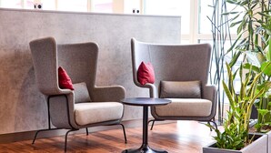 Twee grijze fauteuils met rode kussens, een klein rond tafeltje, en een potplant in een moderne kamer met houten vloer en grote ramen.