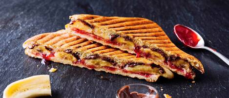 Sándwich a la plancha con relleno visible, una cuchara con mermelada y un tazón pequeño de crema de chocolate sobre una superficie oscura.