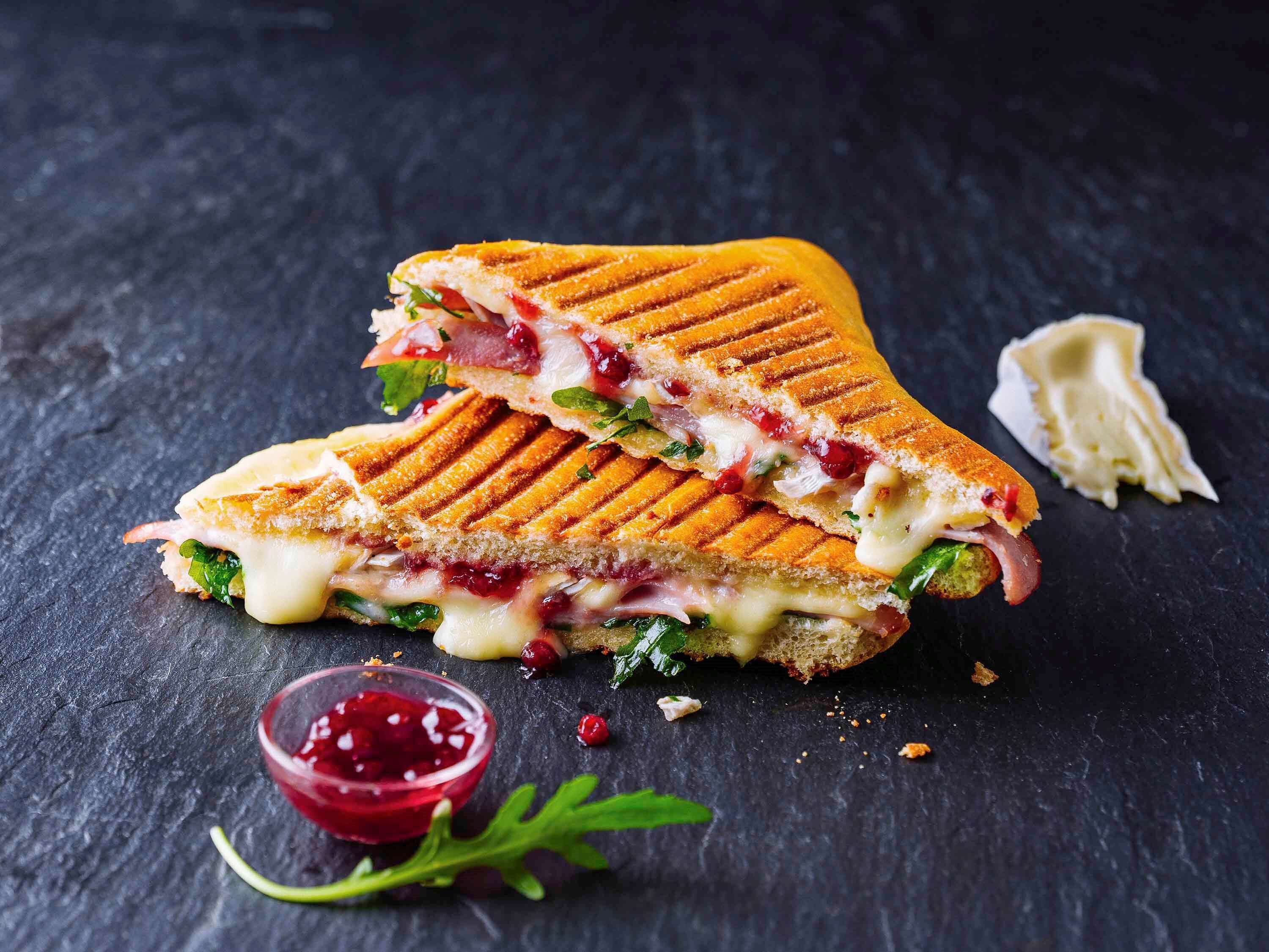 A grilled sandwich with visible layers of ingredients, placed on a dark surface with a small bowl of red condiment and some greens.