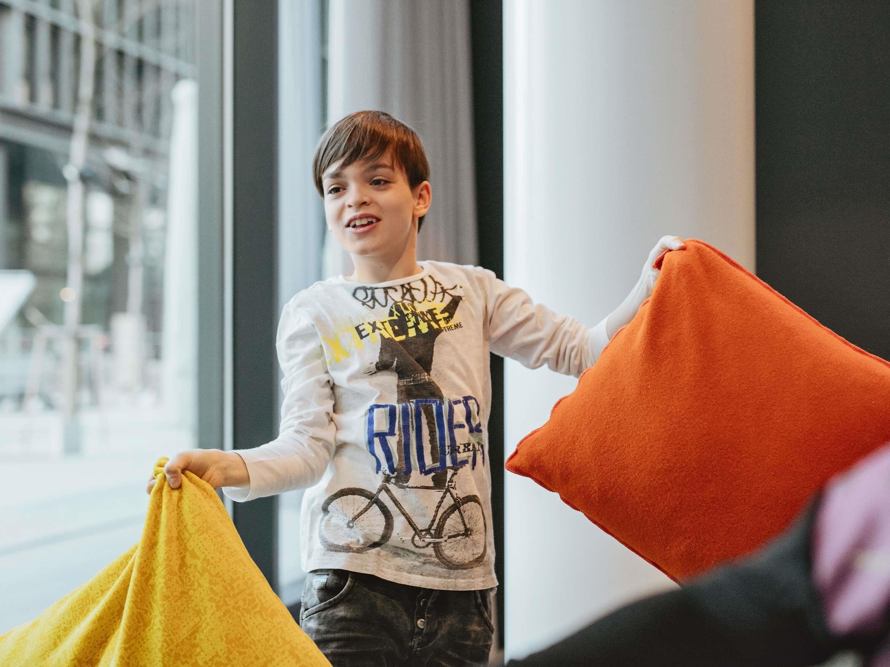 Een kind houdt een gele en oranje kussen vast, staand in een kamer met grote ramen en een zichtbare balustrade.
