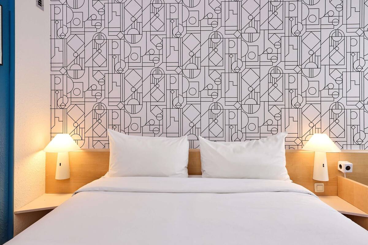 Una habitación de hotel con papel tapiz de diseño geométrico, una cama bien hecha con ropa blanca y dos lámparas de noche.