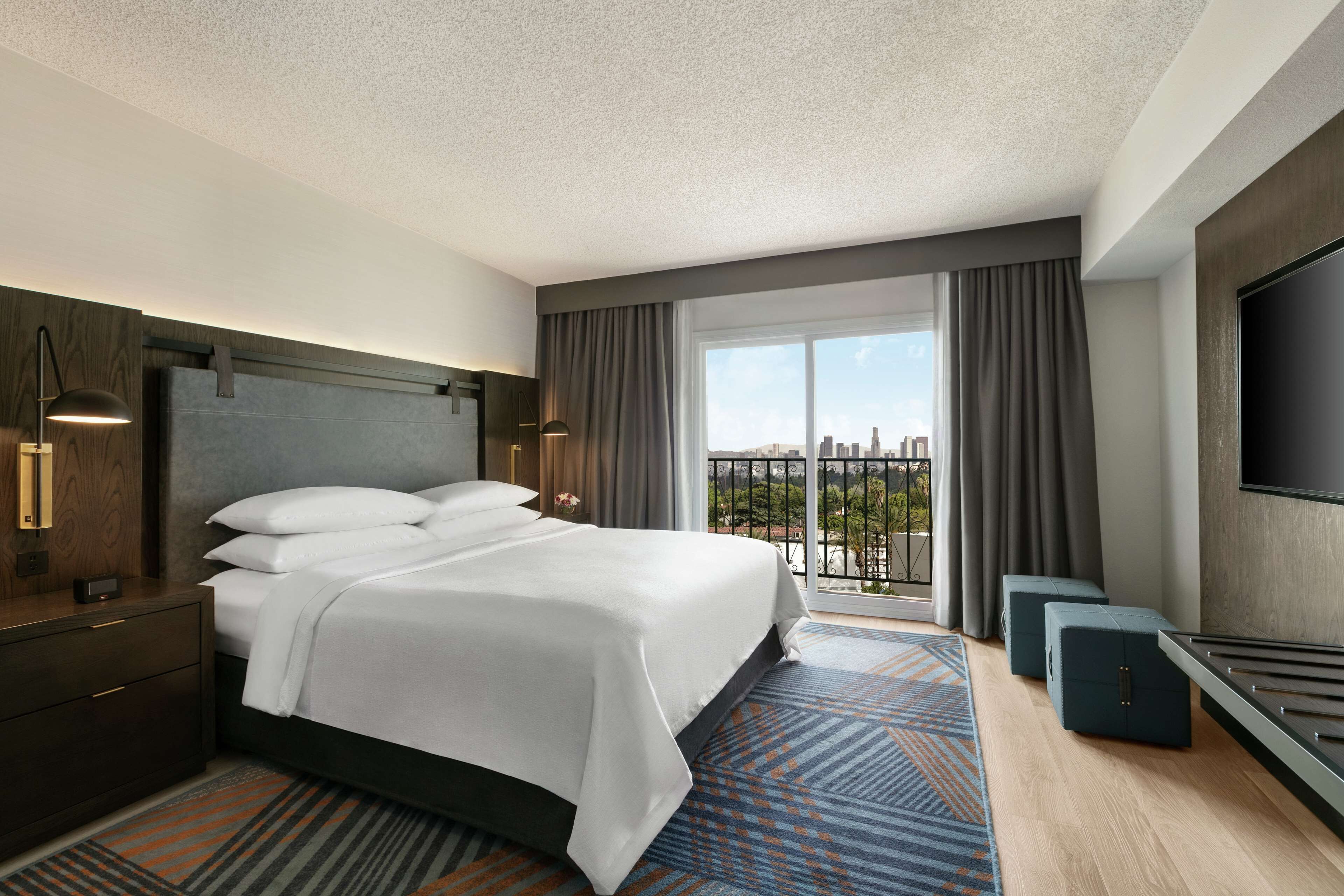 A hotel room with a large bed, a TV, a desk, and a balcony with a city view.