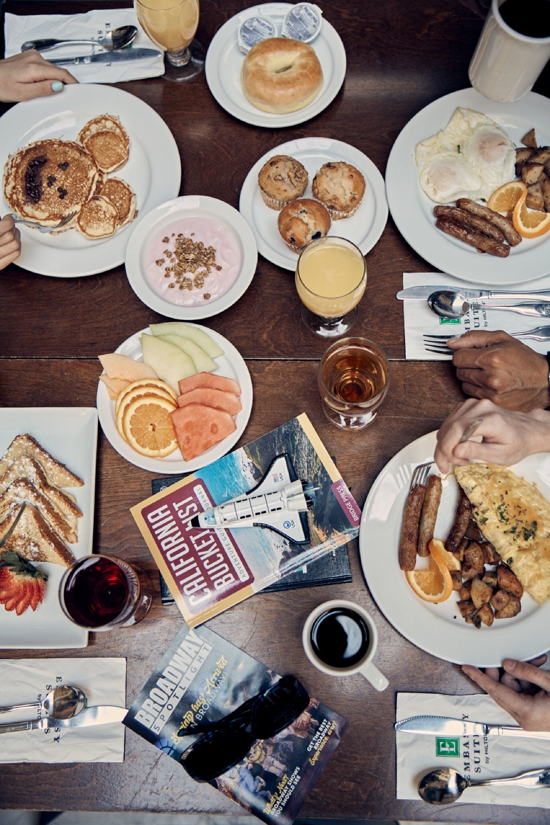 Una mesa con alimentos para el desayuno, como panqueques, huevos, salchichas y tostadas, además de bebidas y una revista.