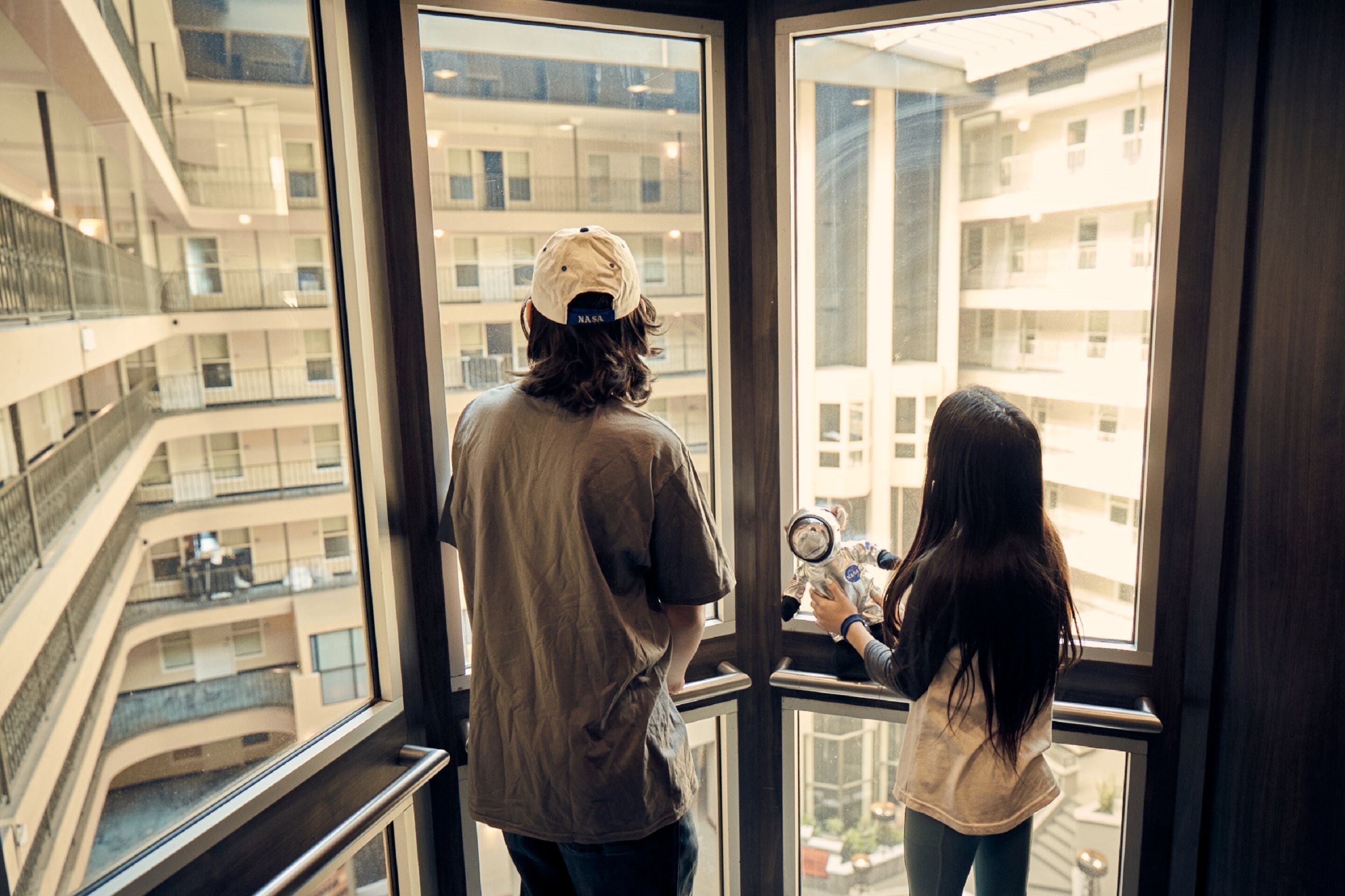 A man and a woman looking out of a large window at a multi-story building with balconies.
