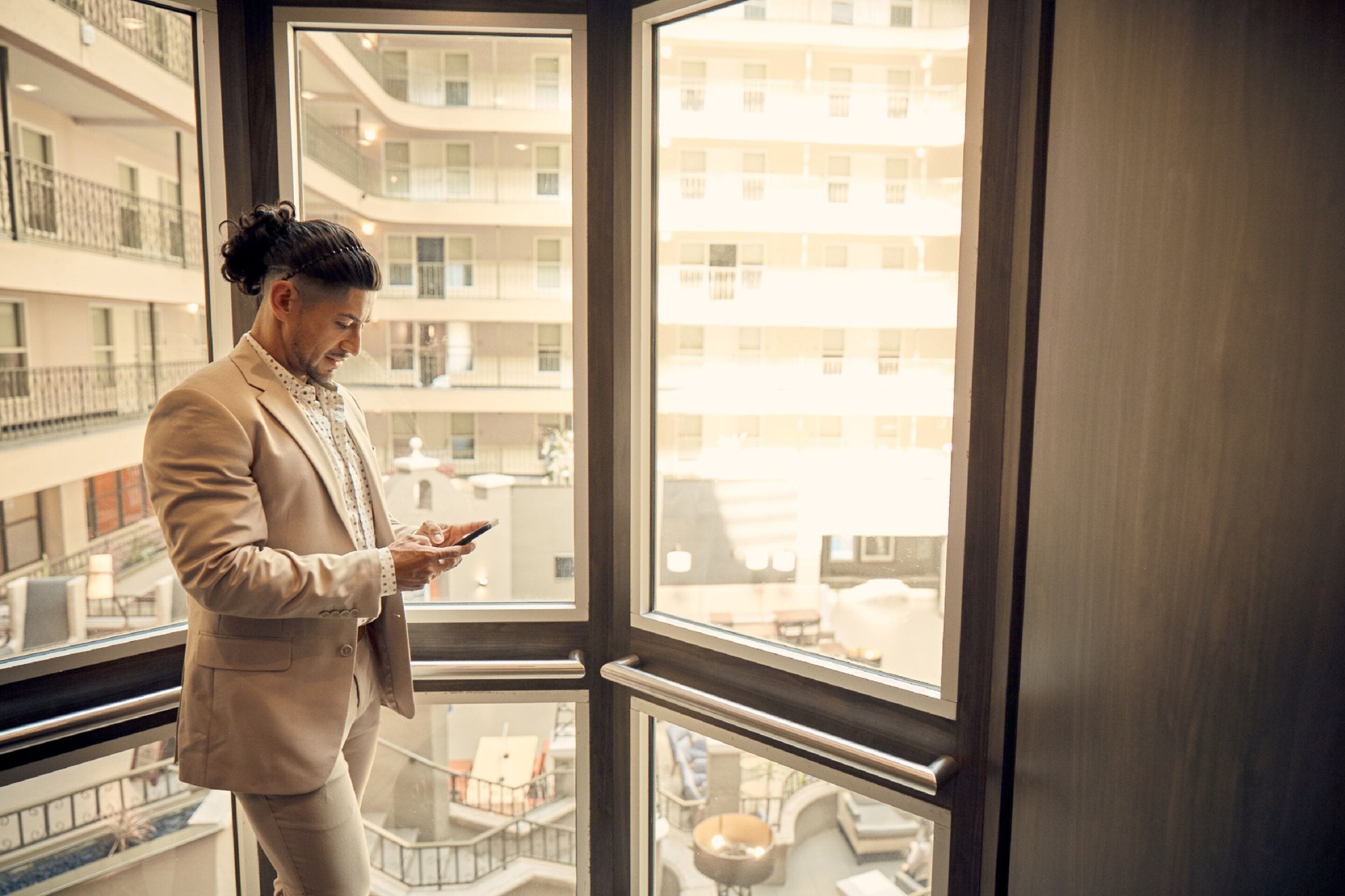 A man in a beige suit standing by a large window with a view of a multi-story building.
