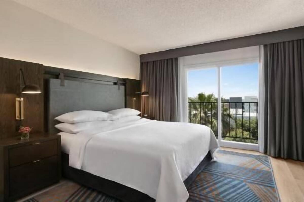 A hotel room with a bed, a nightstand, a lamp, a window with curtains, and a view of the outdoors.