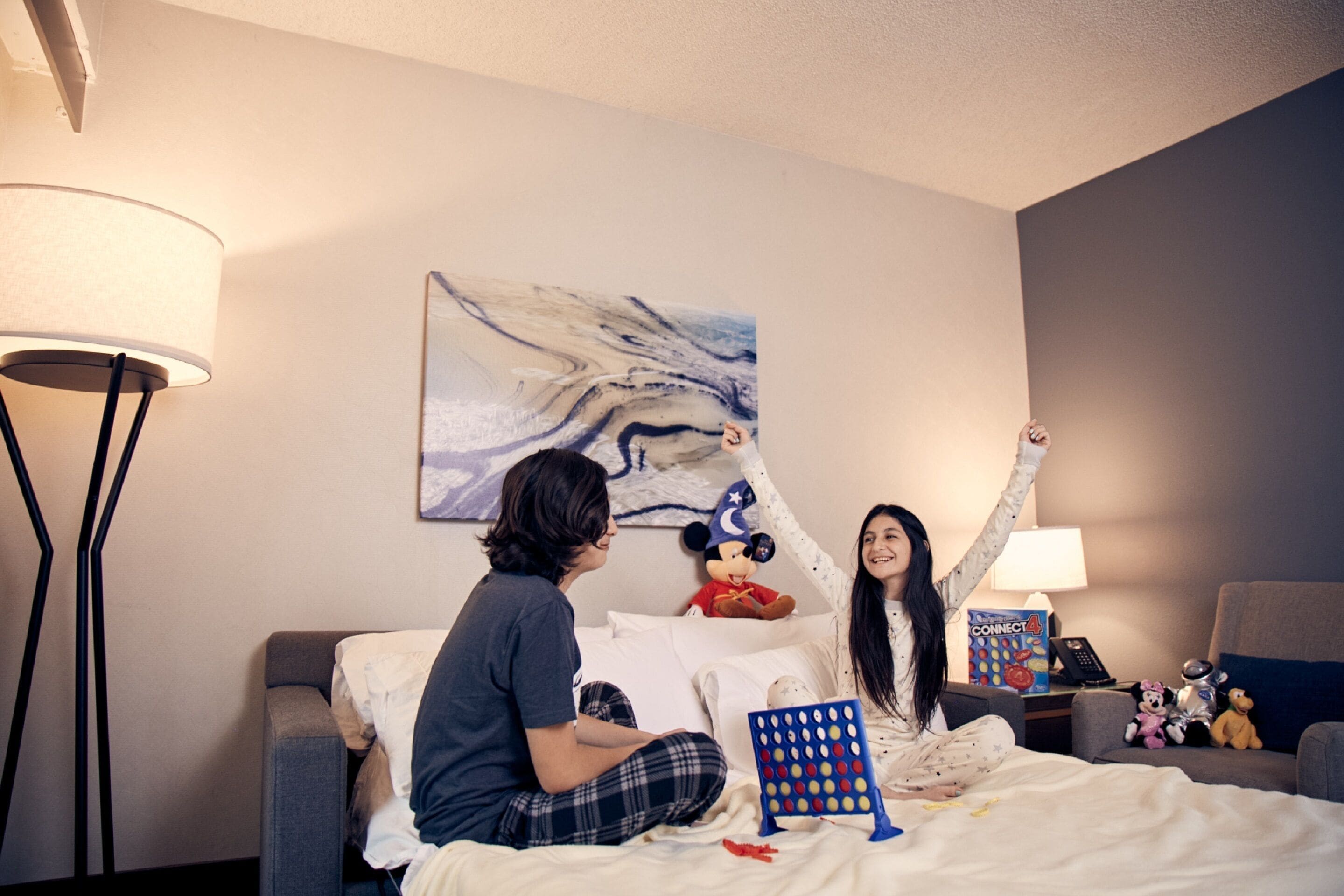 Une chambre d’hôtel avec un lit, une lampe de chevet, un jouet en peluche à l’effigie de Mickey Mouse et un jeu de société Connect 4.