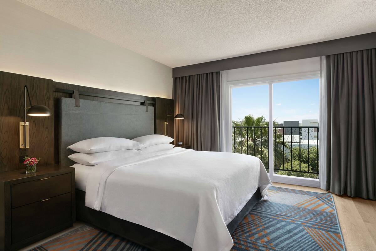 A hotel room with a large bed, a nightstand with a lamp, a view of the outdoors through a balcony, and a window with curtains.