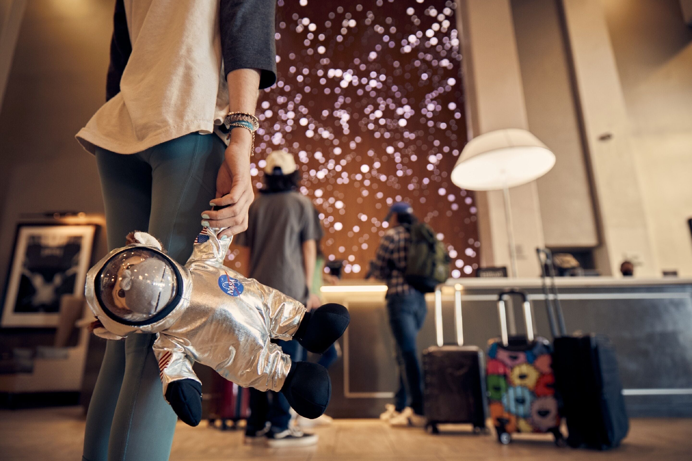 A person holding a child in a space suit in a lobby with a reception desk and luggage.