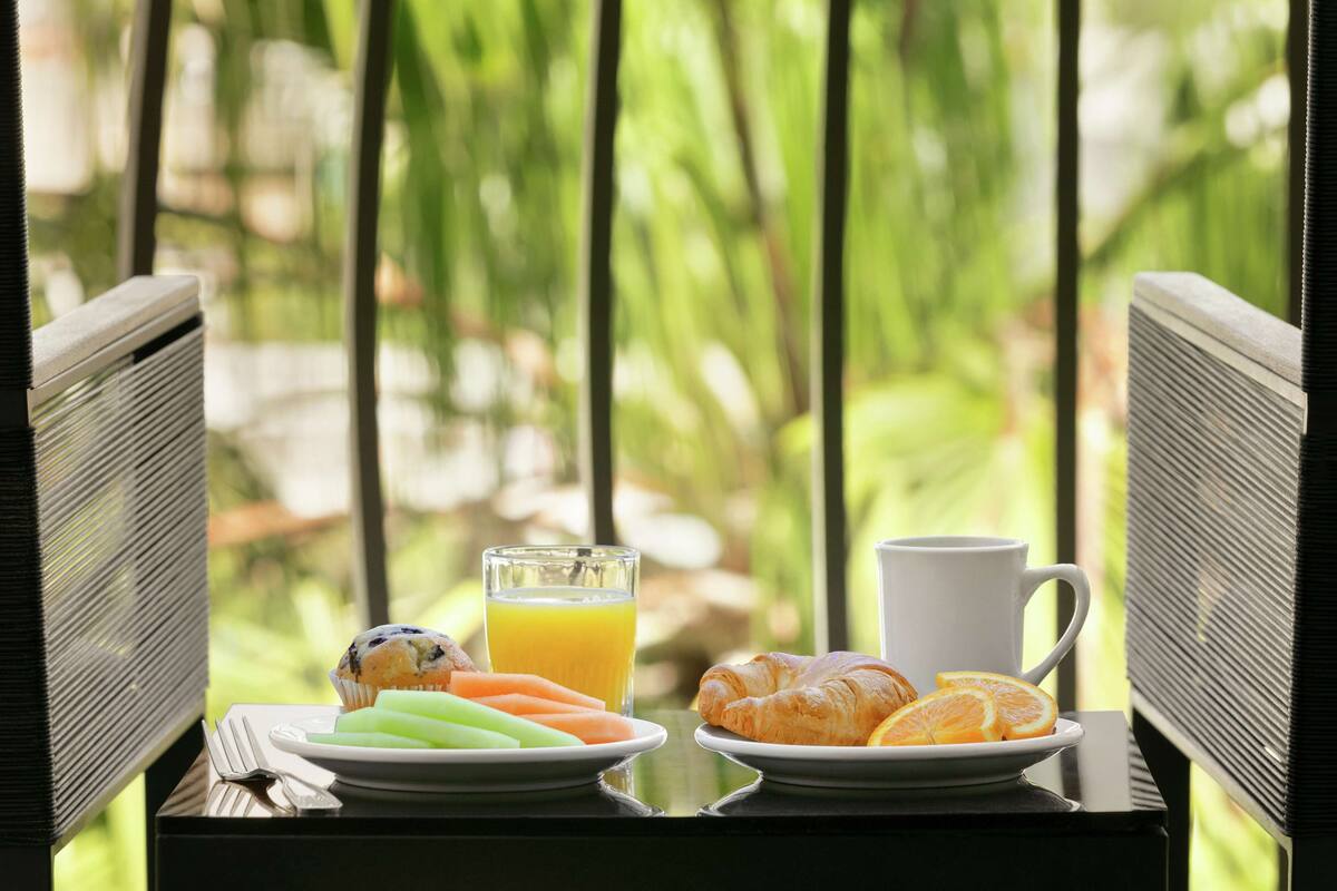 A breakfast setup with plates of food and drinks on a table, set against a backdrop of greenery.