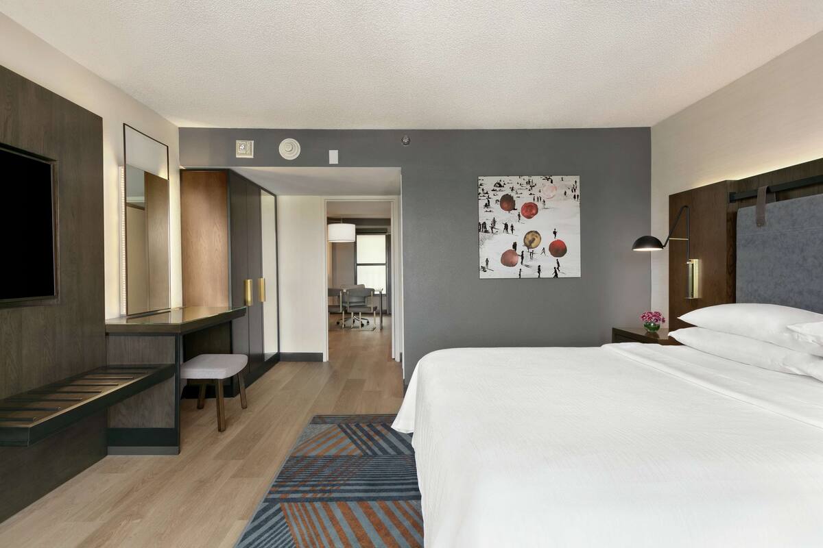 A modern hotel room with a large bed, a desk with a chair, a large mirror, and a wall art piece.