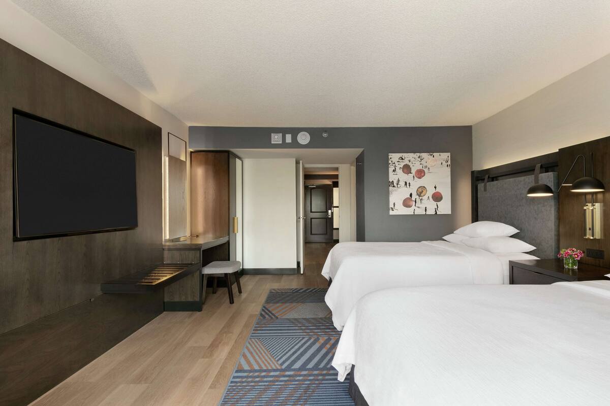 A hotel room with two beds, a desk, a flat-screen TV, and a painting on the wall.