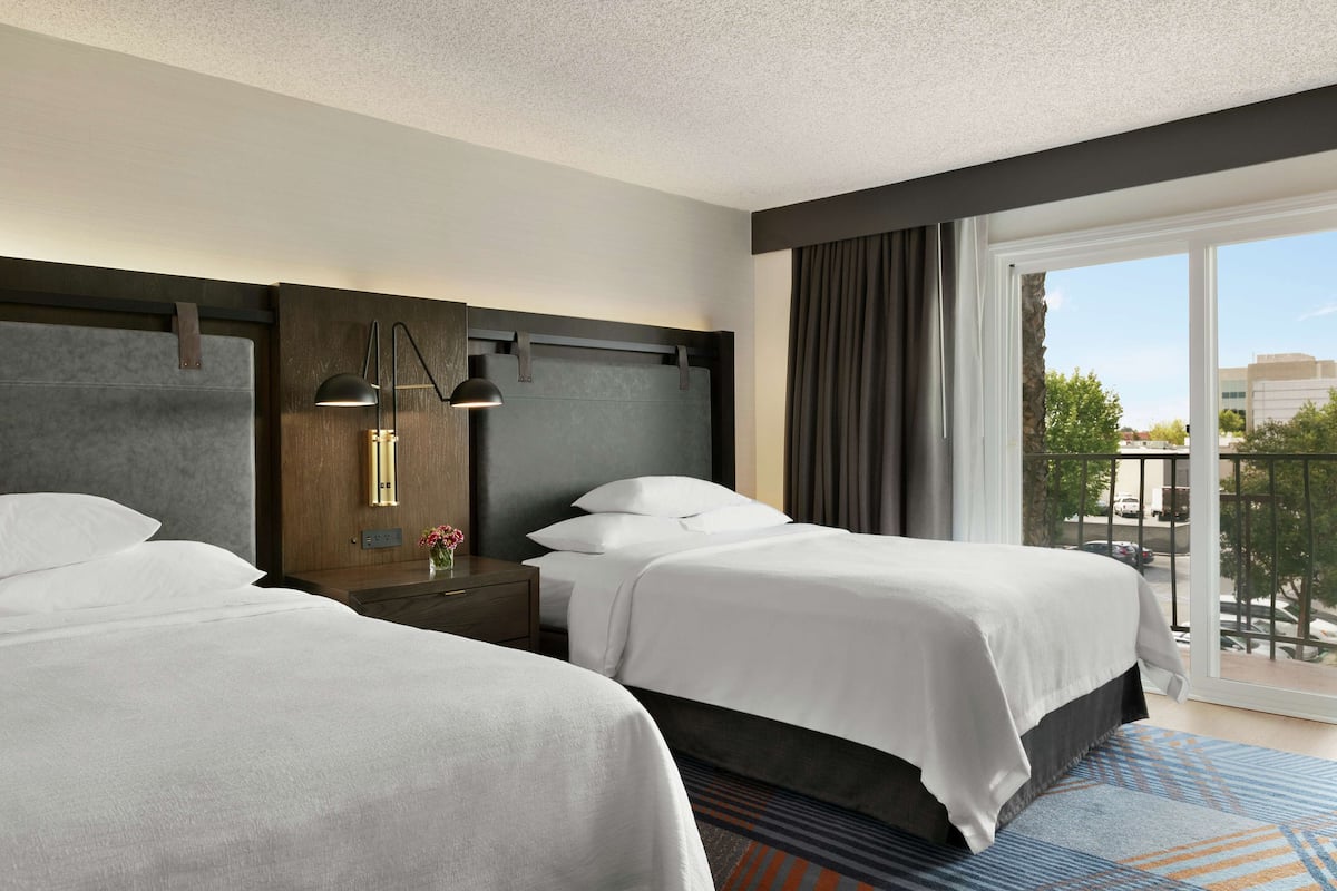 A hotel room with two beds, a nightstand with a lamp, a view of the outdoors through a large window, and a balcony.