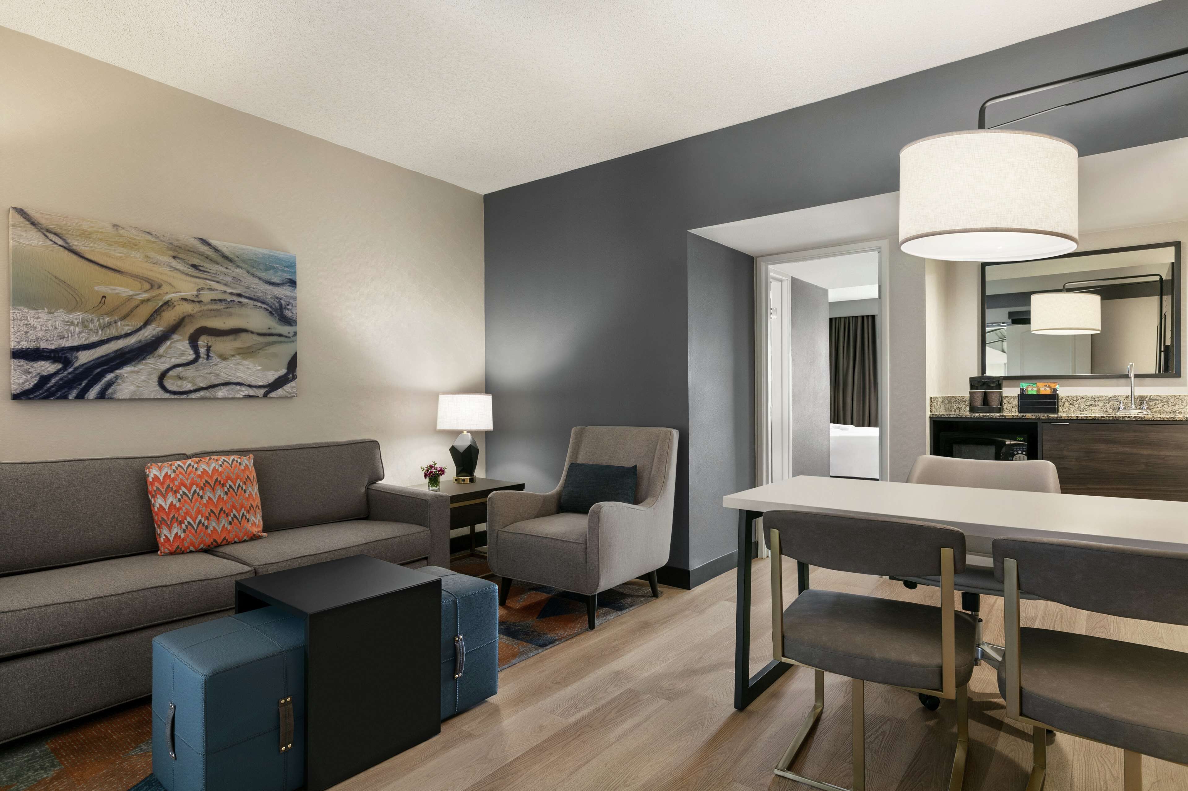 A modern living room with a gray sofa, a blue ottoman, a gray armchair, and a dining table with chairs. There is a painting on the wall, a lamp on a side table, and a kitchen area visible through an open doorway.