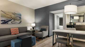 A modern living room with a gray sofa, a blue ottoman, a gray armchair, and a dining table with chairs. There is a painting on the wall, a lamp on a side table, and a kitchen area visible through an open doorway.