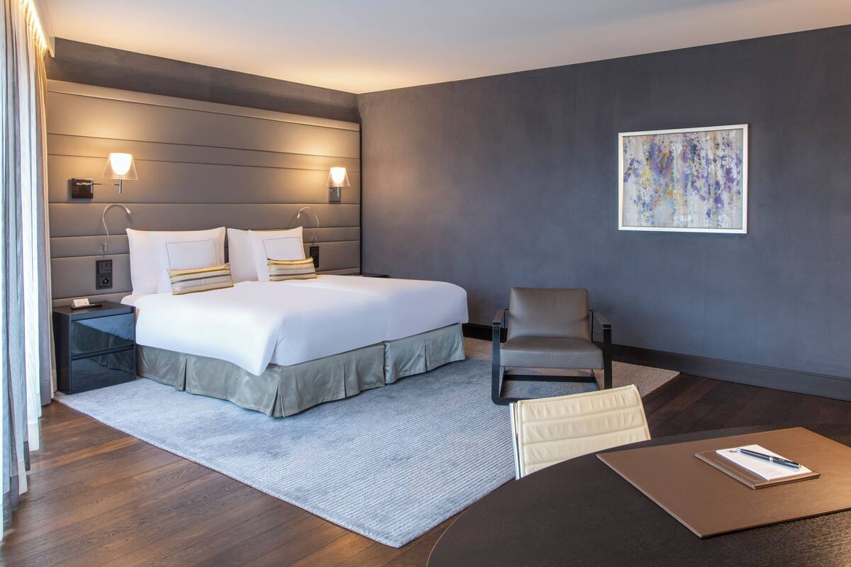 Una habitación de hotel moderna con una cama grande, lámparas de noche, una silla, un escritorio y una obra de arte enmarcada en la pared.