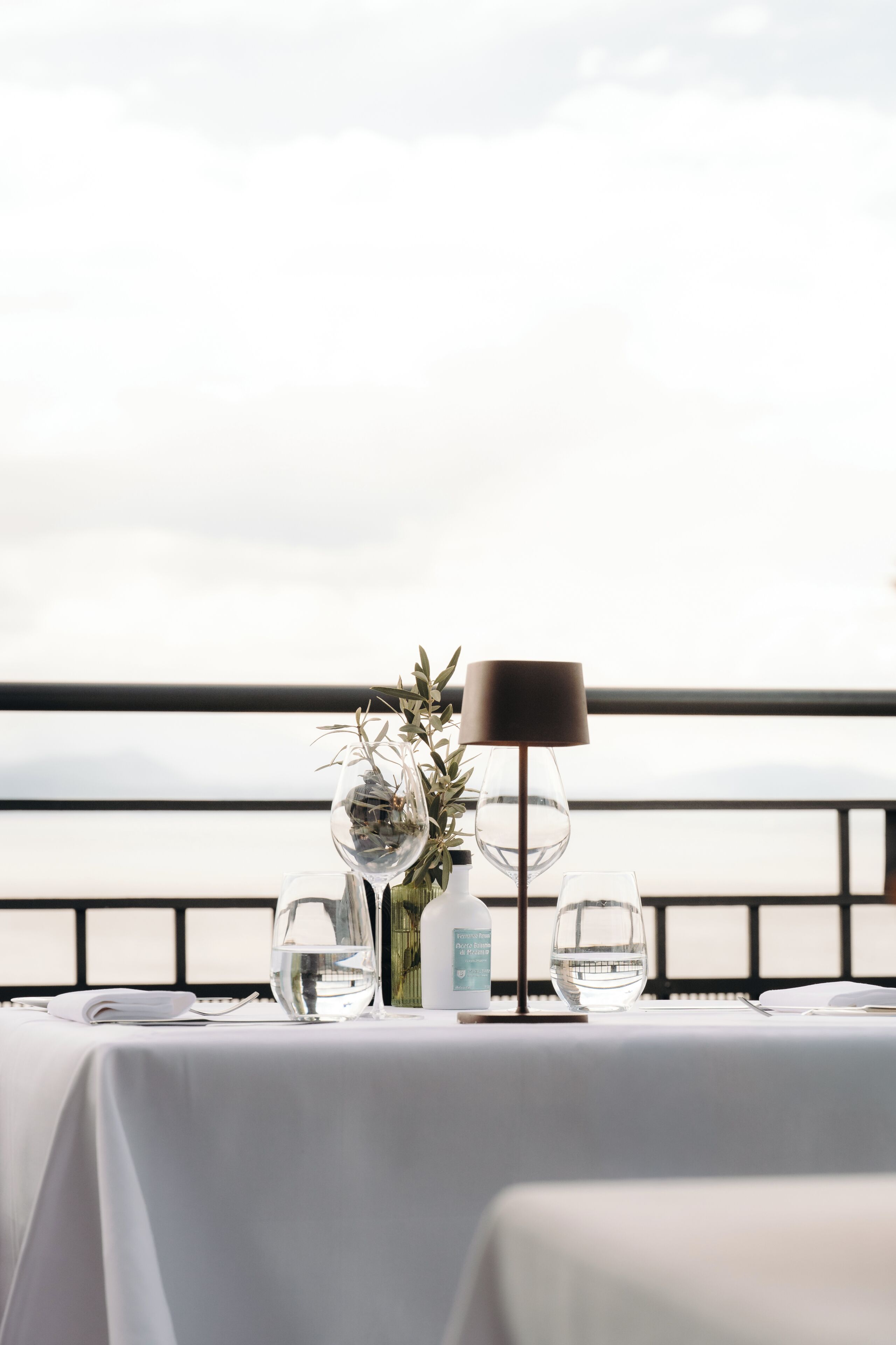 A table set with a water bottle, glasses, and a vase with greenery, overlooking a body of water.