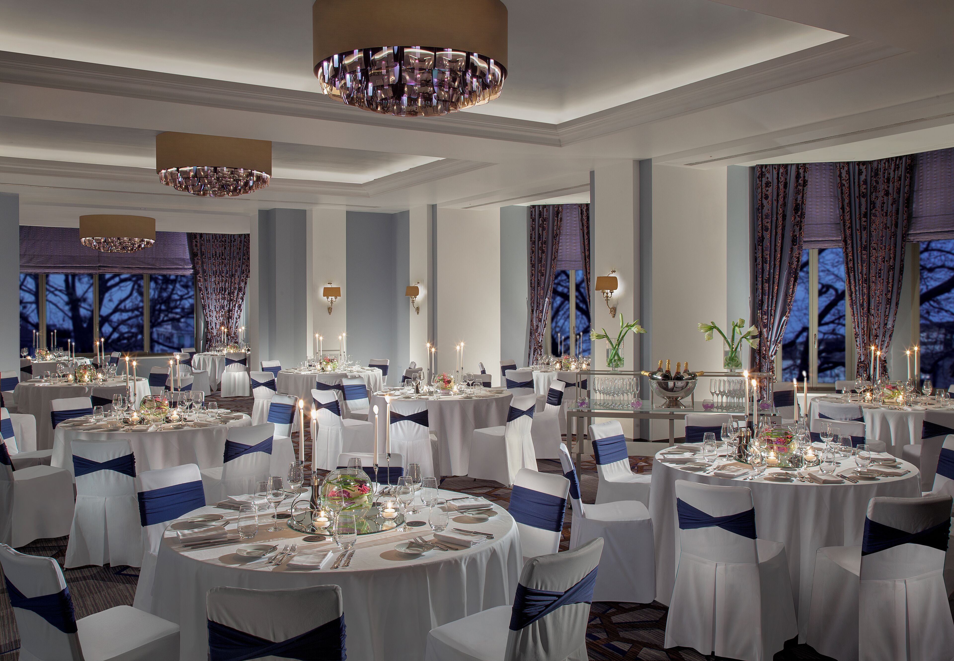 A banquet hall with round tables set for a formal event, featuring white tablecloths, blue sashes, and candlelit centerpieces.