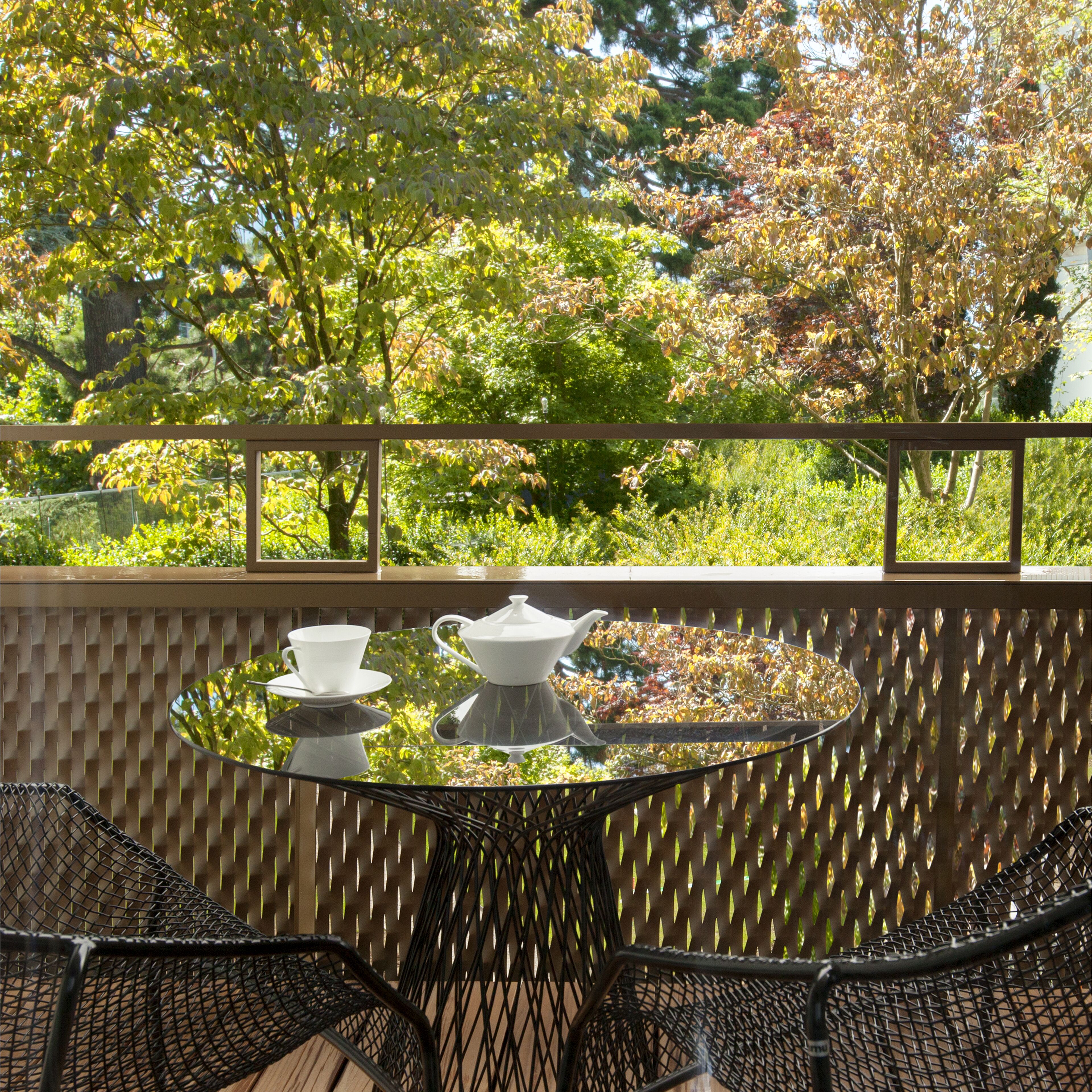 A round table with a white teapot and cup, set against a lattice wall with a view of trees and foliage.