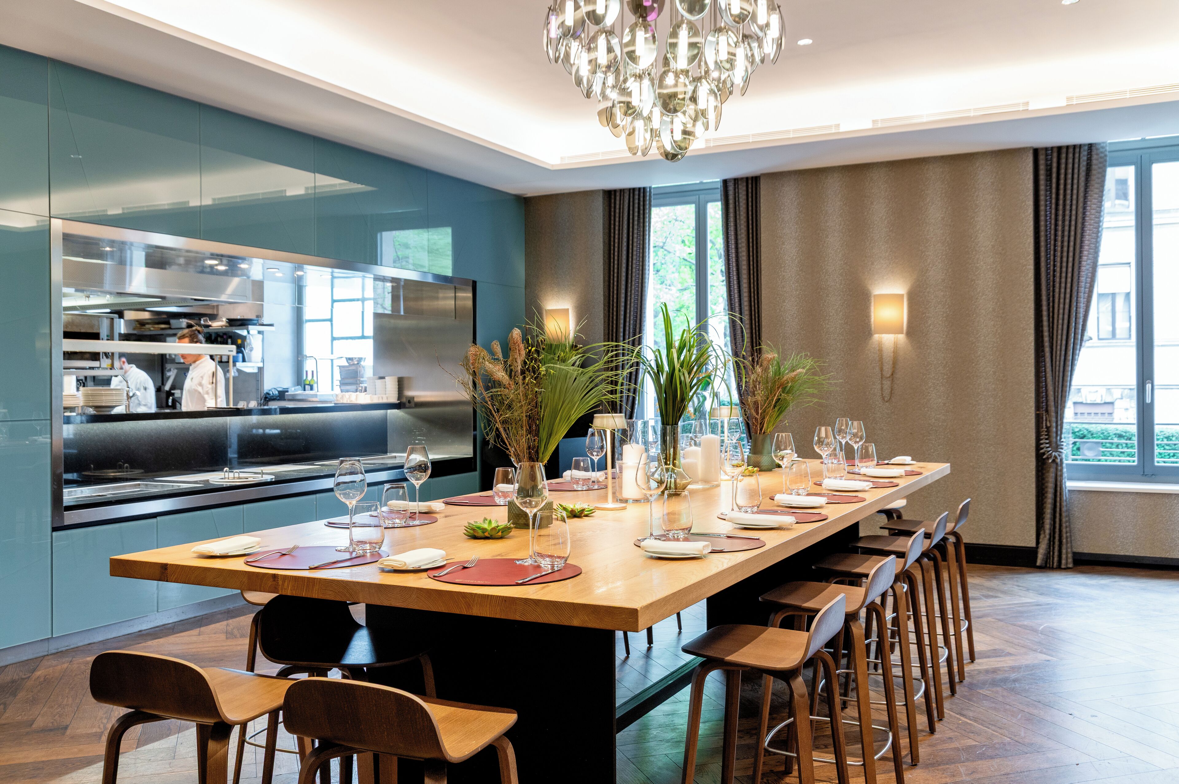 Un comedor moderno con una mesa larga dispuesta para una comida, una cocina al fondo y un amplio ventanal con cortinas.