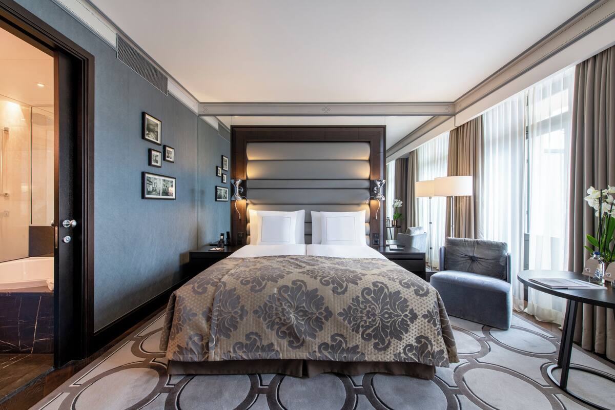 Una habitación de hotel con una cama grande, mesitas de noche, una silla y una pequeña mesa con lámpara.