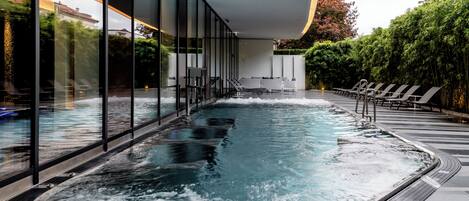 Una piscina interior moderna con paredes de vidrio, sillas de descanso y una cascada.