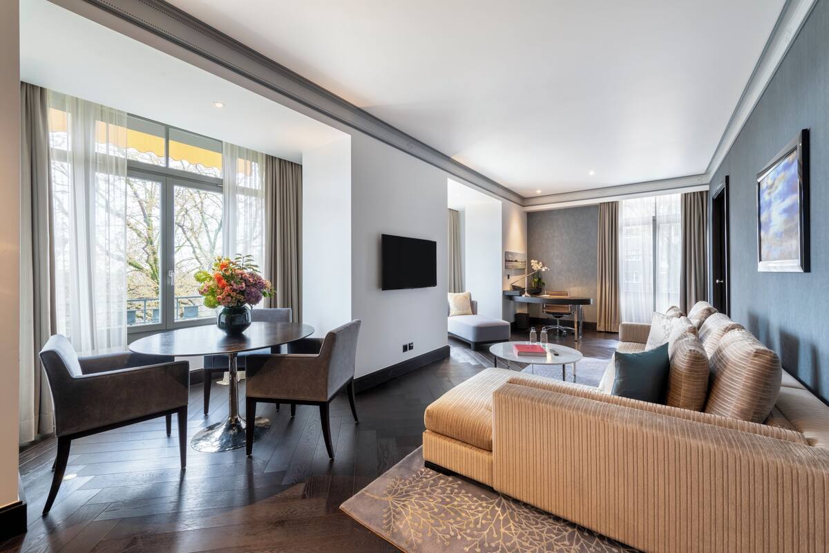 Una sala de estar moderna con un sofá, una mesa de comedor y sillas, un televisor de pantalla plana montado en la pared y ventanas amplias.