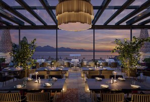 Een terras met tafels en stoelen, een grote hanglamp en uitzicht op bergen en zee.