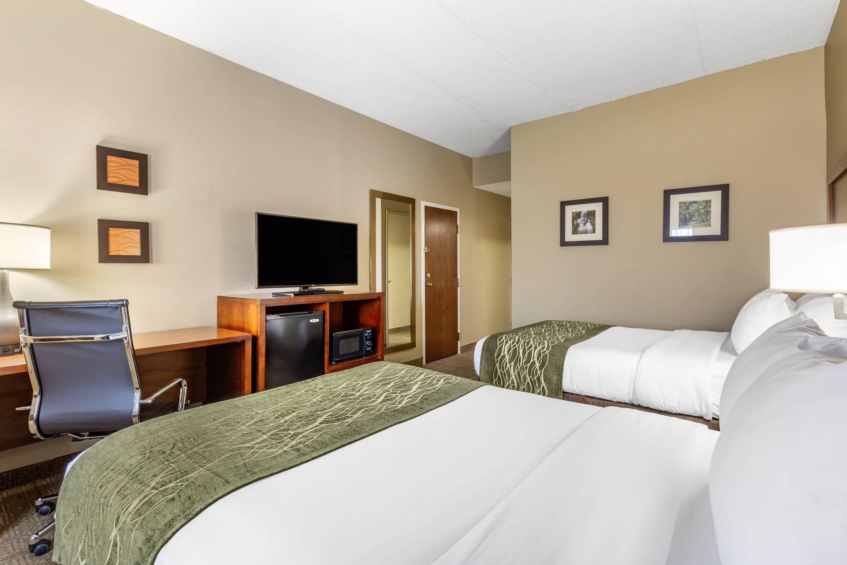 Una habitación de hotel con dos camas, un escritorio con una computadora, un televisor y una puerta.